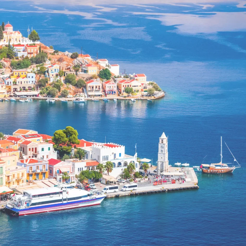 Πανοραμική άποψη παραθαλάσσιου οικισμού σε ελληνικό νησί με πολύχρωμα παραδοσιακά σπίτια και γραφικό λιμάνι.