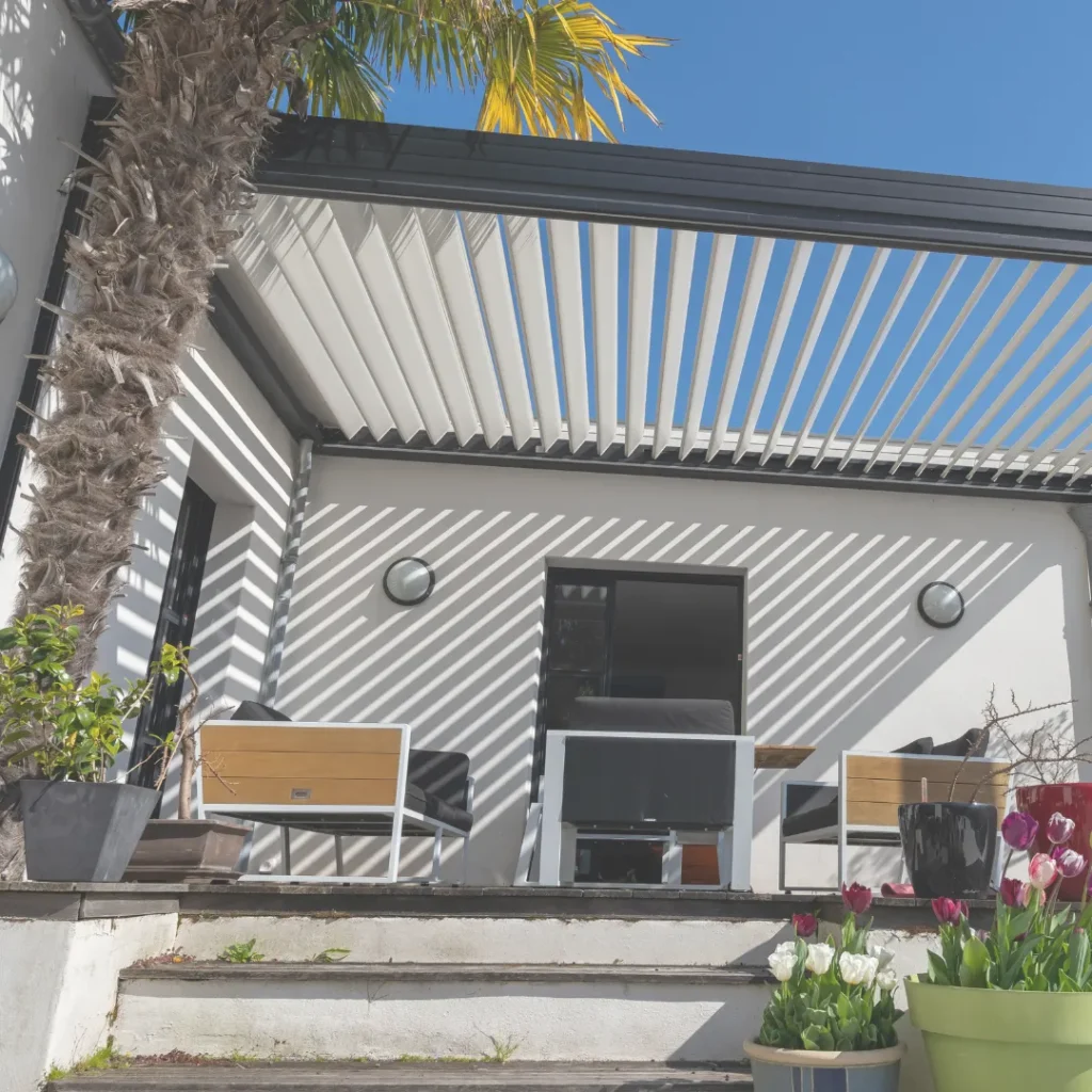 Μοντέρνα βιοκλιματική πέργκολα με περσίδες που δημιουργεί ρίγες σκιάς σε αυλή σπιτιού, ένα παράδειγμα έξυπνης αρχιτεκτονικής σκίασης.